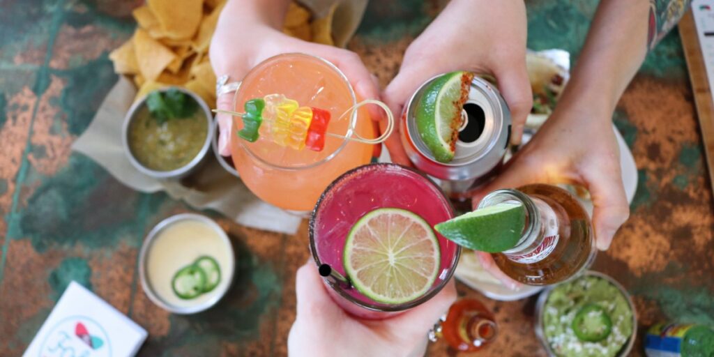 Hands holding various colorful alcoholic beverages topped with food